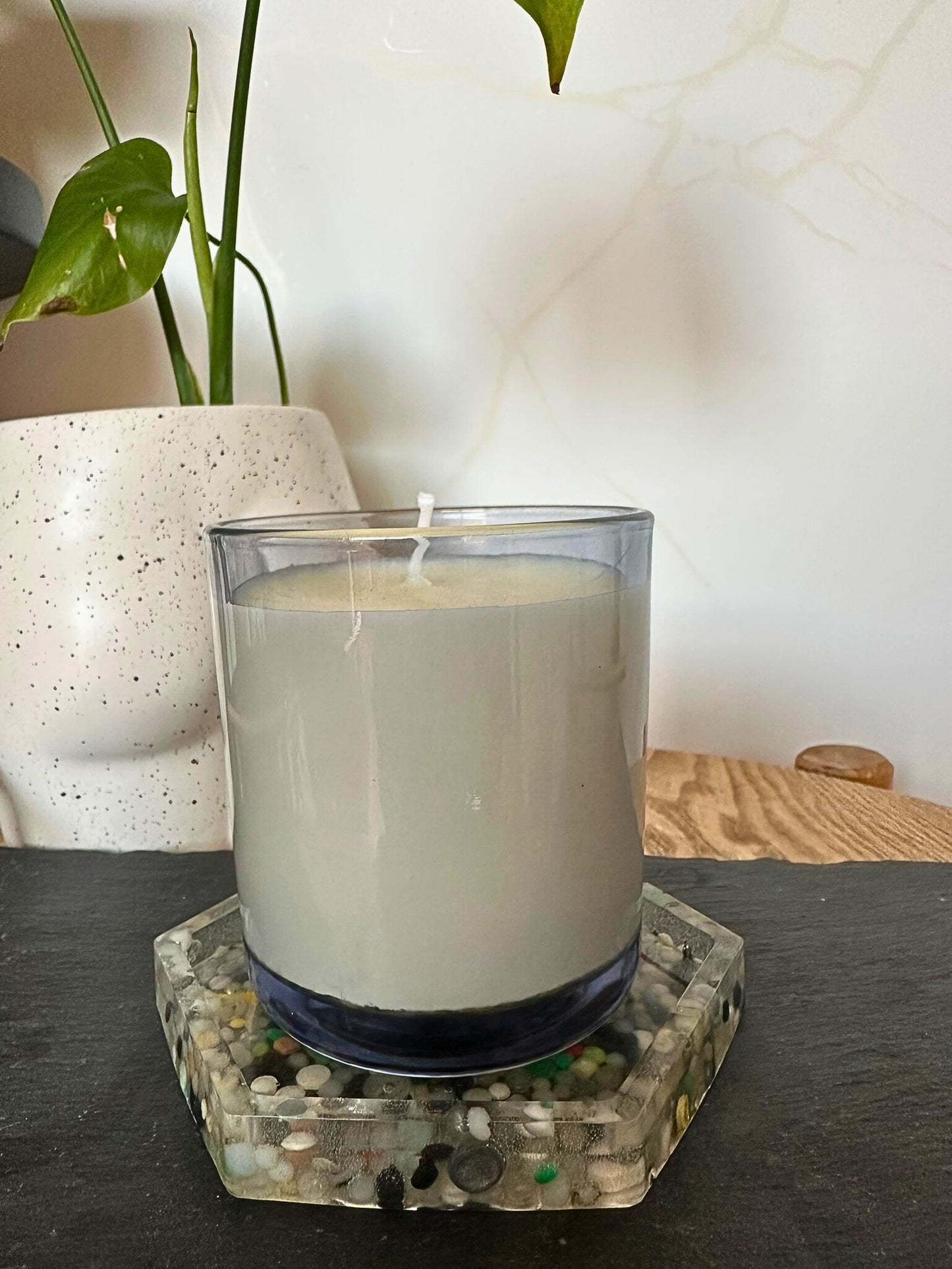 White candle in a glass holder on a decorative coaster with a plant in the background.