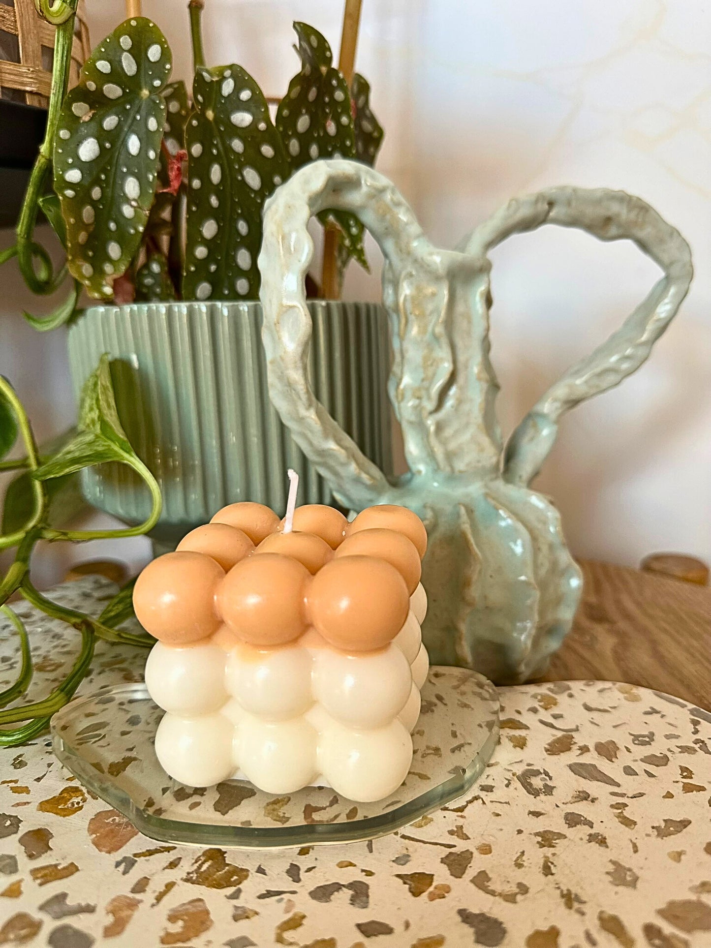 Cylindrical candle with spherical design on a decorative plate, with plants and a ceramic vase in the background.