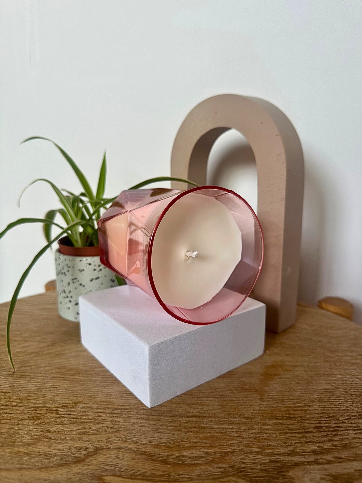 Decorative Suki Home The Pink Hikari Candle on a white base, placed on a wooden surface with a plant in the background.