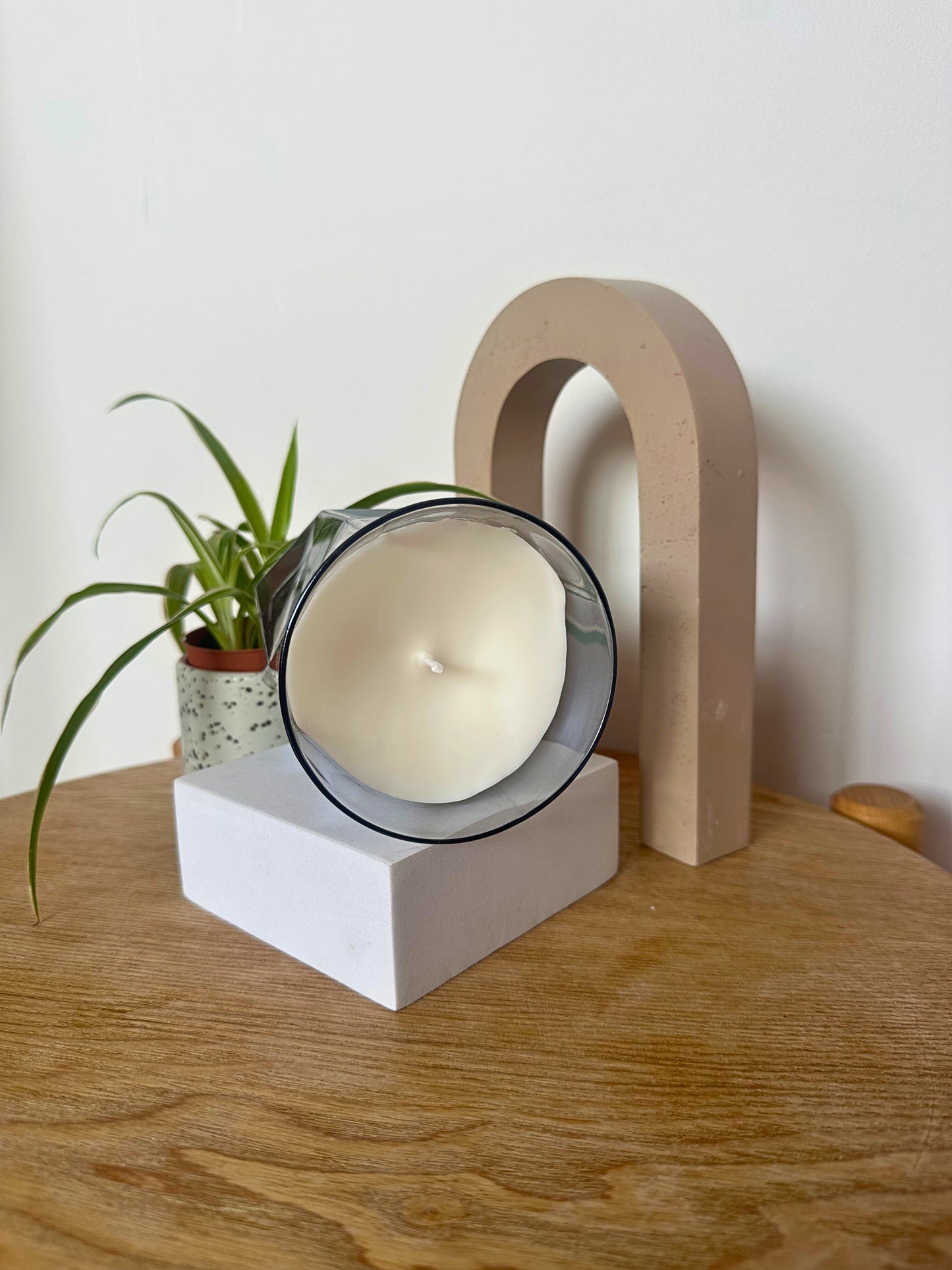 The Hikari Candle in Grey by Suki Home on a wooden surface with a plant and wooden arch in the background