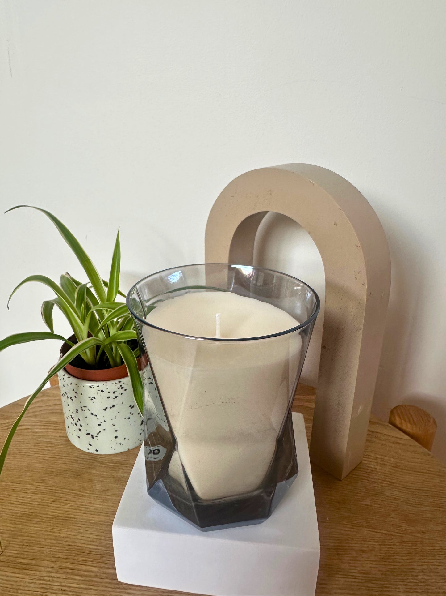 The Hikari Candle in Grey by Suki Home on a wooden surface with a plant and decorative item in the background.
