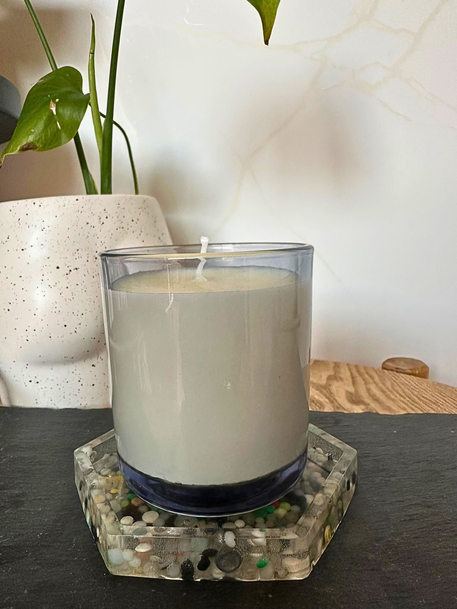 White candle in a glass holder on a decorative coaster with a plant in the background.