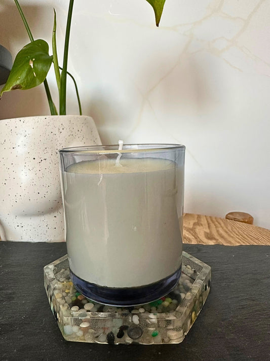 White candle in a glass holder on a decorative coaster with a plant in the background.