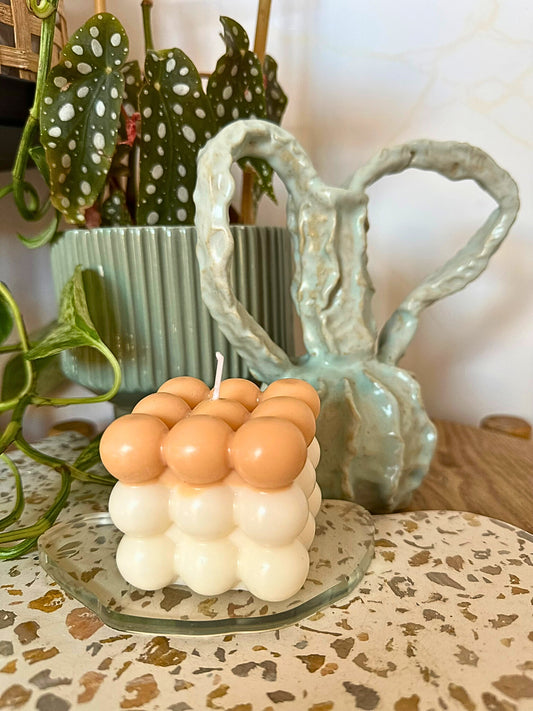 Cylindrical candle with spherical design on a decorative plate, with plants and a ceramic vase in the background.