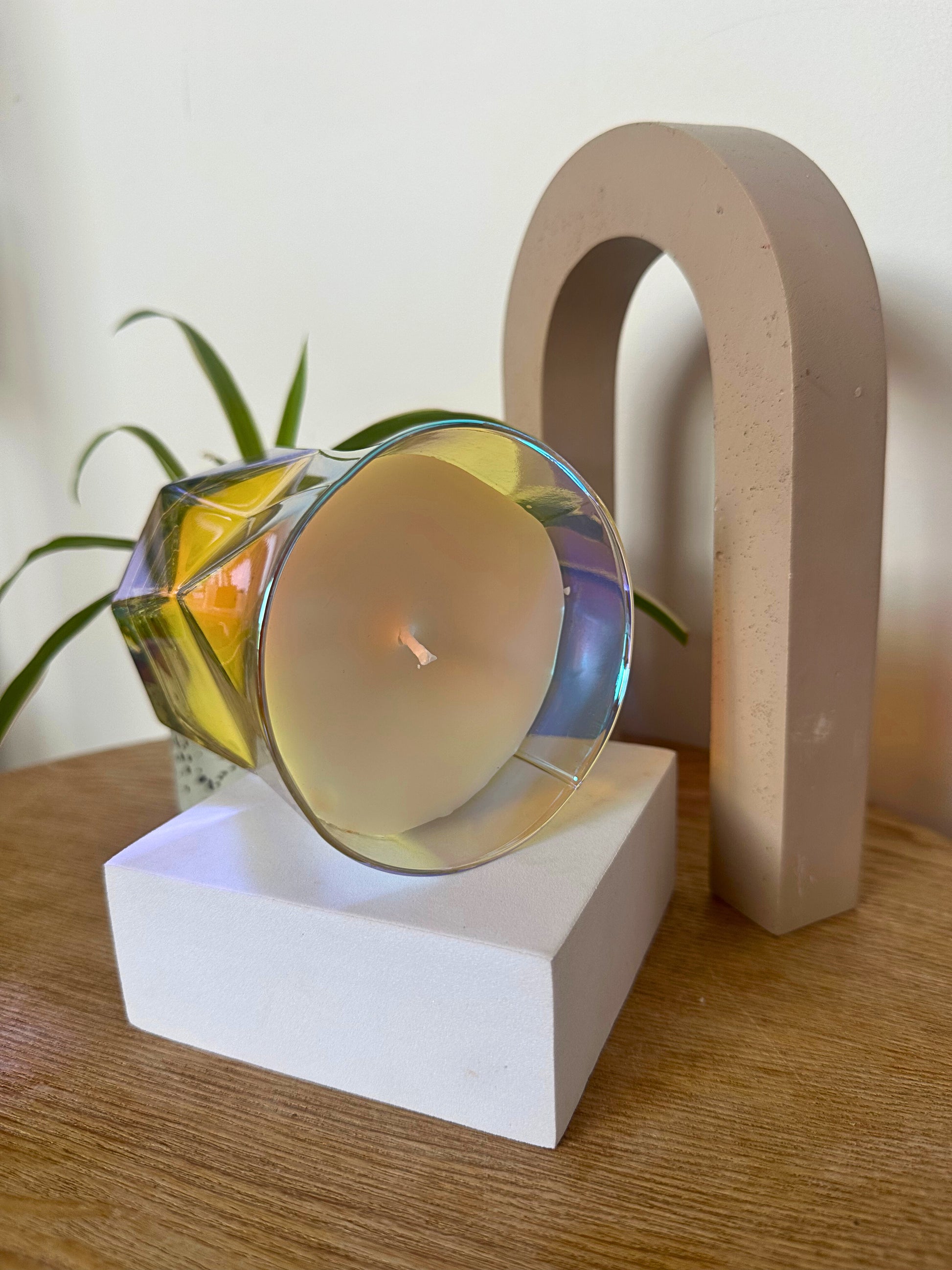 Suki Home The Hikari Candle in Crystal on side laying on a white base with a plant and wooden object in the background.
