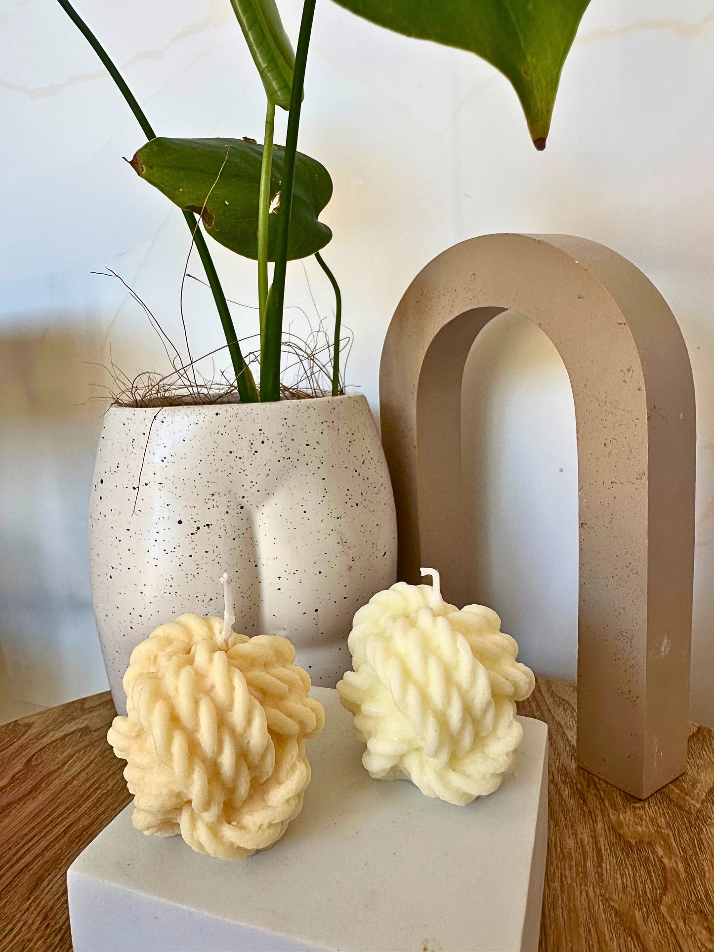 Two textured candles on a white block with a plant and decorative arch in the background.