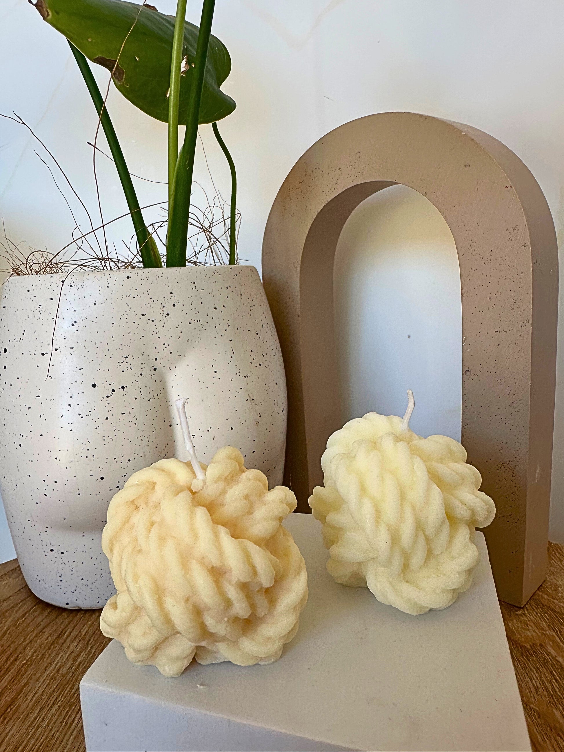 Two textured candles on a white surface with a plant and decorative arch in the background.