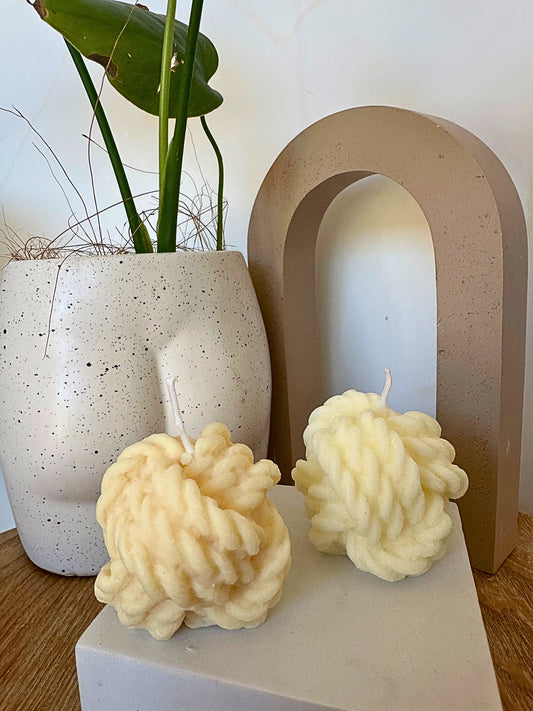 Two textured candles on a white surface with a plant and decorative arch in the background.