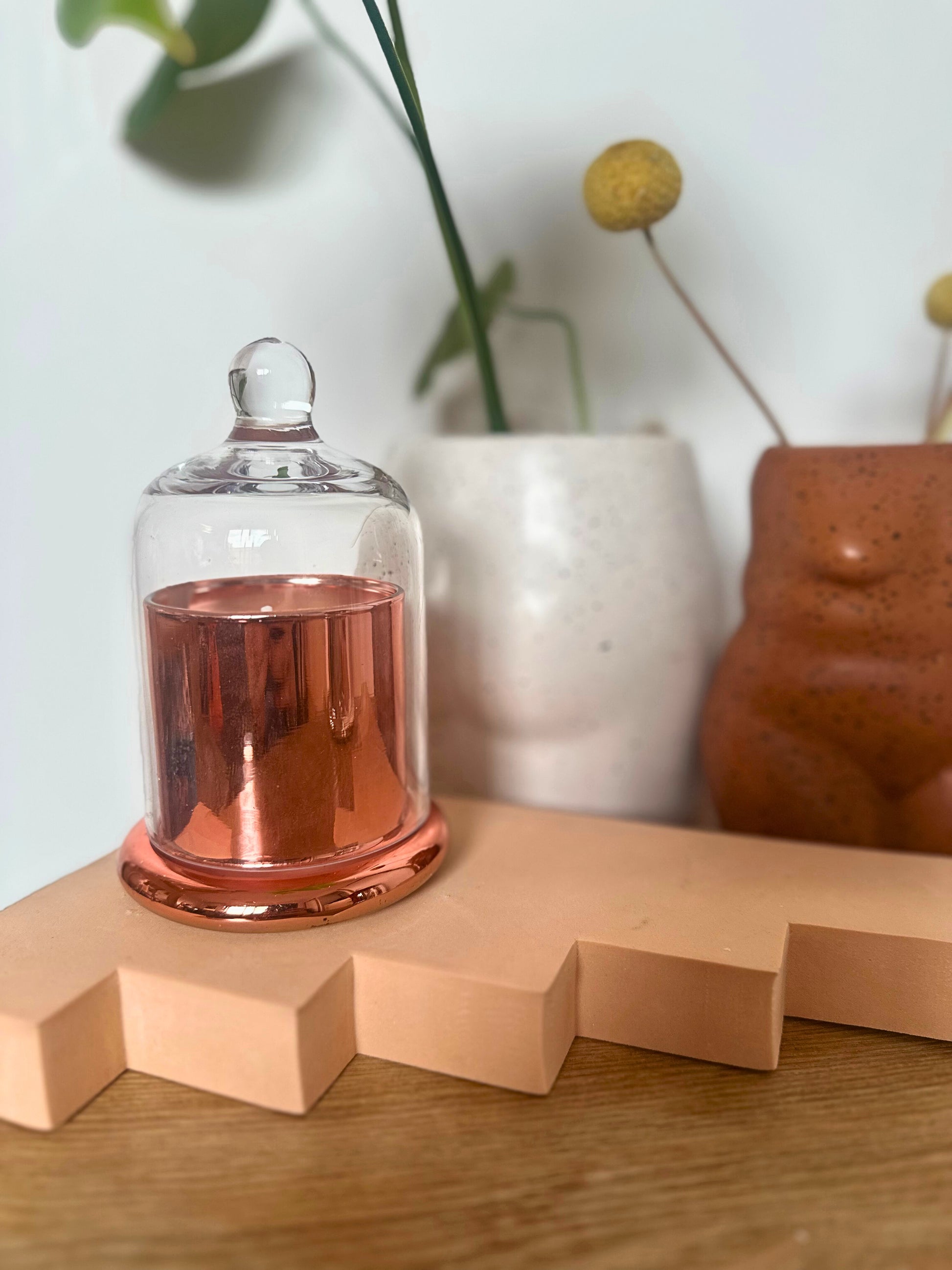 Suki Home The Seina Cloche Candle in Gold on a wooden surface with decorative vases in the background.