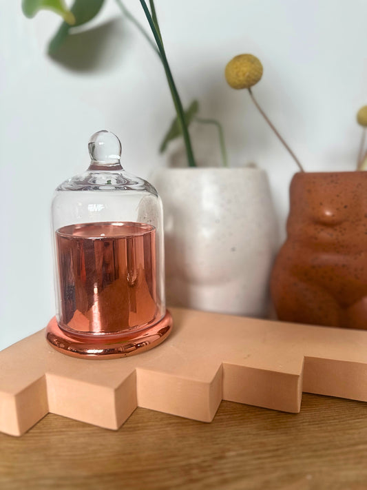 Suki Home The Seina Cloche Candle in Gold on a wooden surface with decorative vases in the background.