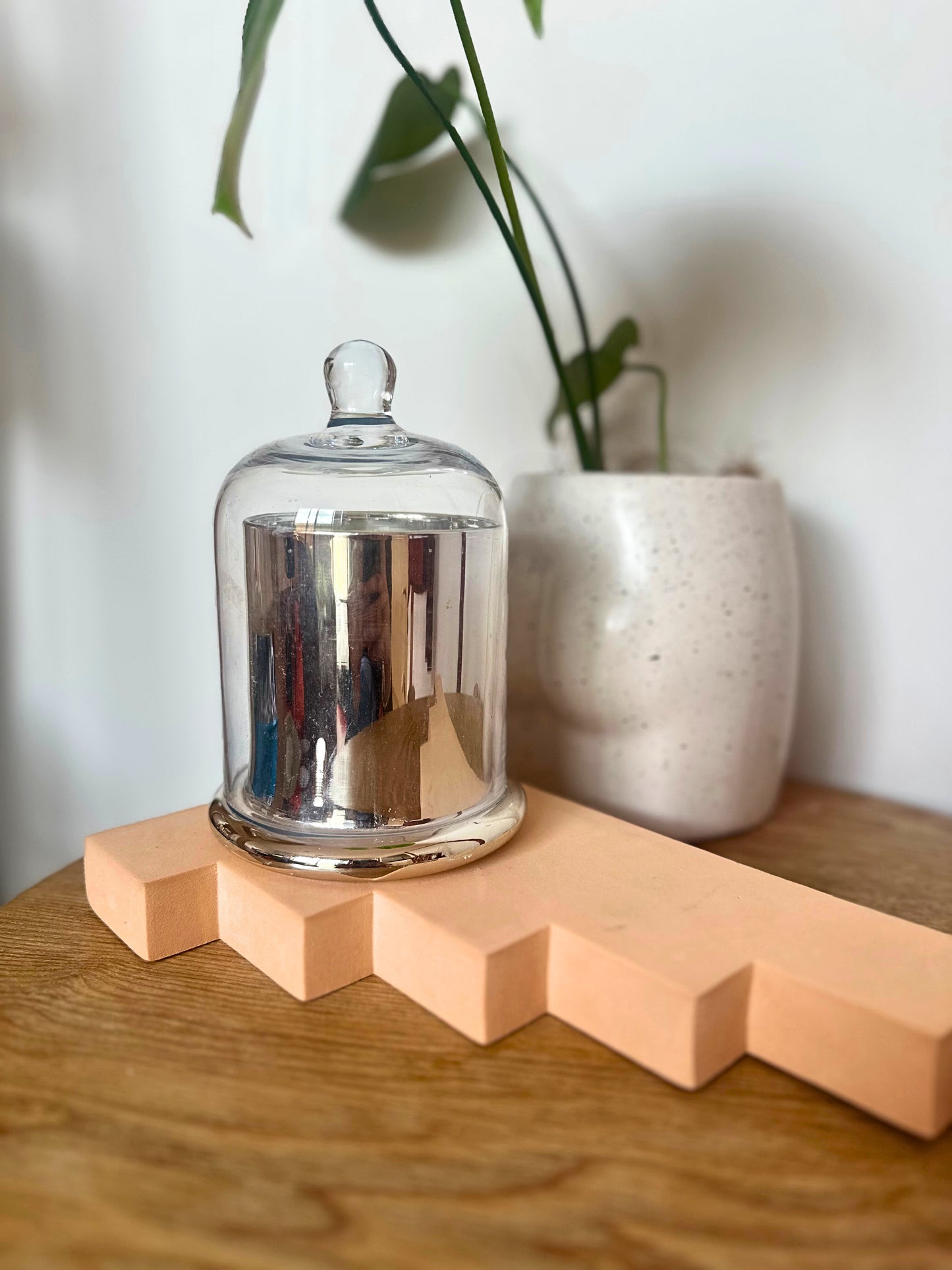 Suki Home The Seina Cloche Candle in Large Silver on peach colored blocks with a white vase and plant in the background.