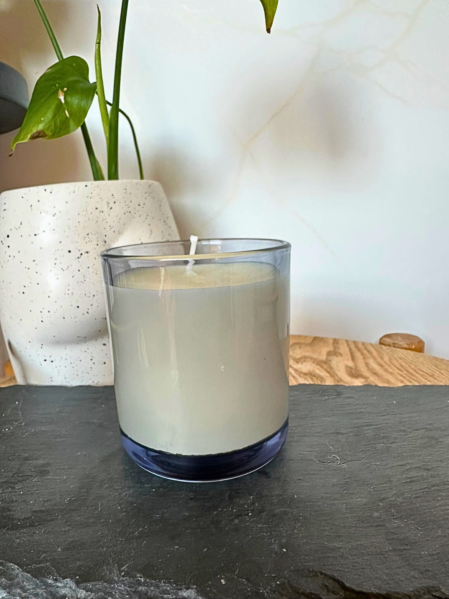 Candle in a glass container on a dark surface with a plant in the background
