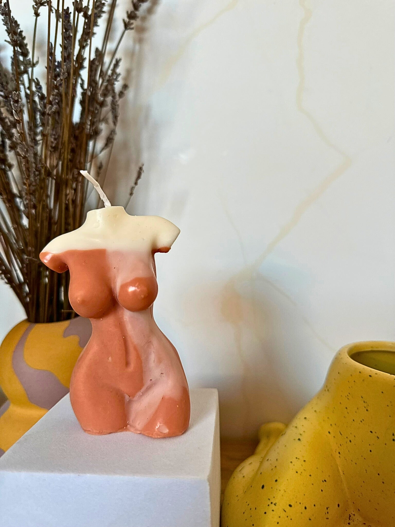 Candle shaped like a female torso with a yellow top and pink body, placed on a white surface with a blurred background.