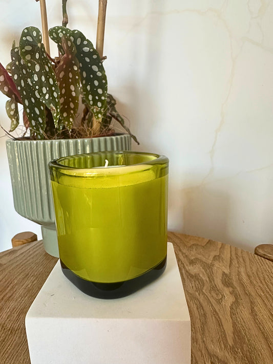 Green candle in a glass container on a wooden surface with a plant in the background