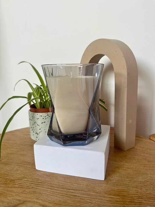 The Hikari Candle in Grey by Suki Home on a white block with a plant and decorative arch in the background.