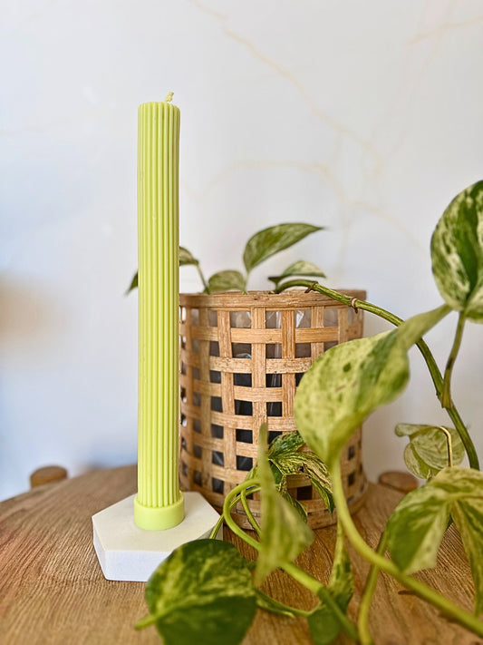 Green candle on a white base with a plant and woven basket in the background